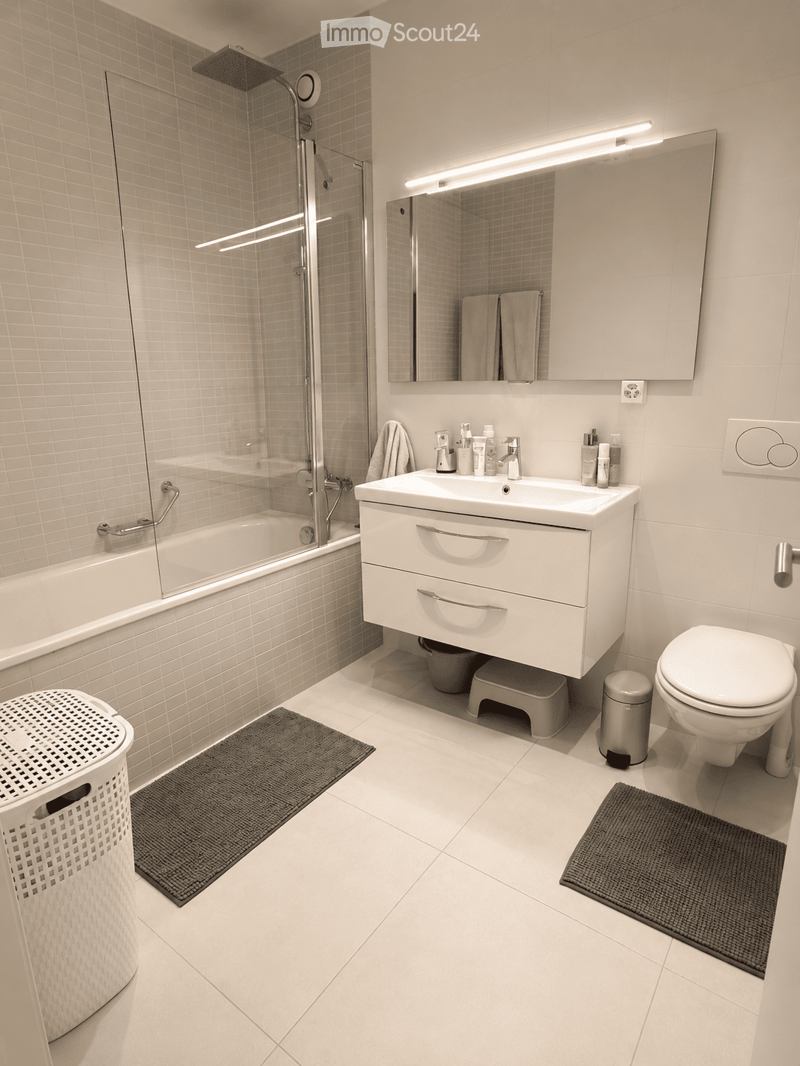 Modern bathroom with white tiles, bathtub, sink, toilet, mirror, and shower. Includes towels, a cabinet, and a trash can.