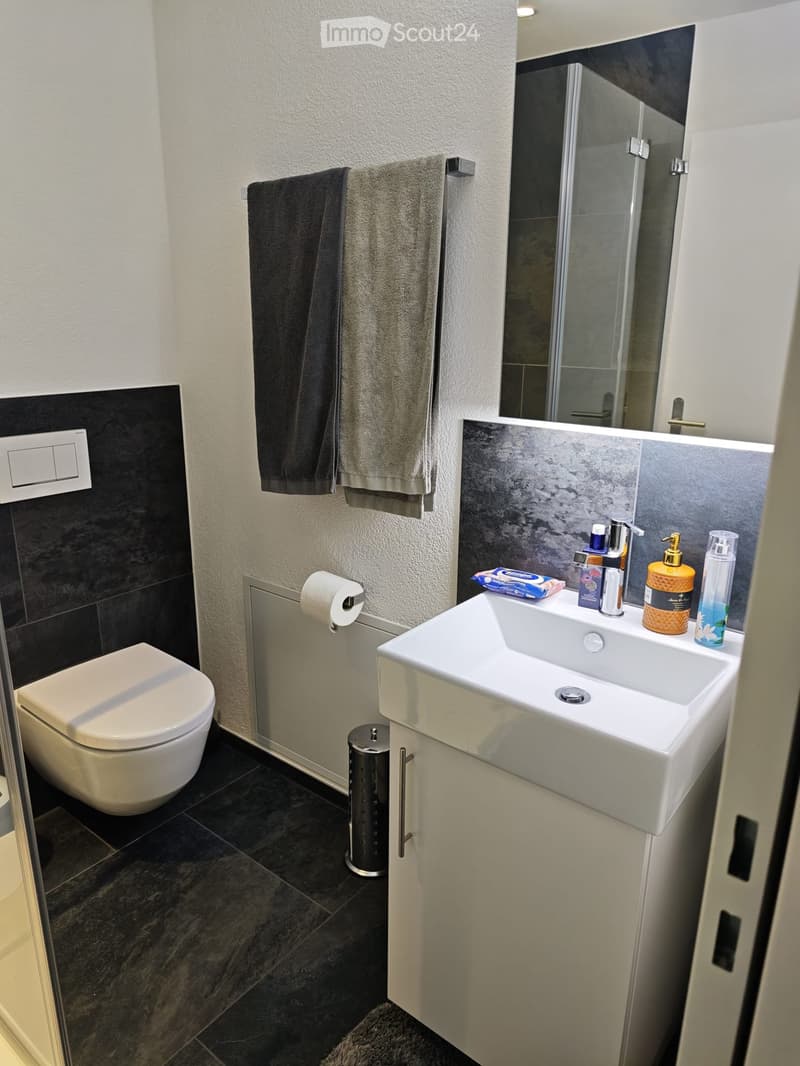 Modern bathroom with a white toilet, a white sink with a modern faucet, a mirror, and a towel hanging on a rack. The sink has various toiletries, and the floor is tiled in dark gray.