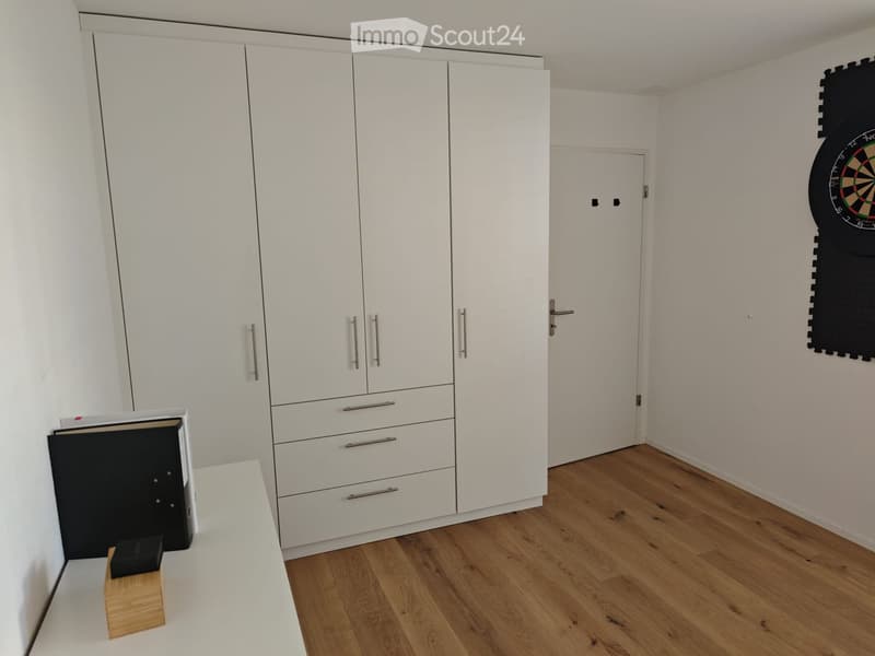 A modern, minimalist room with white walls, wooden flooring, and a large white wardrobe with multiple drawers and doors. A small table and chair are positioned near the wardrobe, and a dartboard is mounted on the wall.