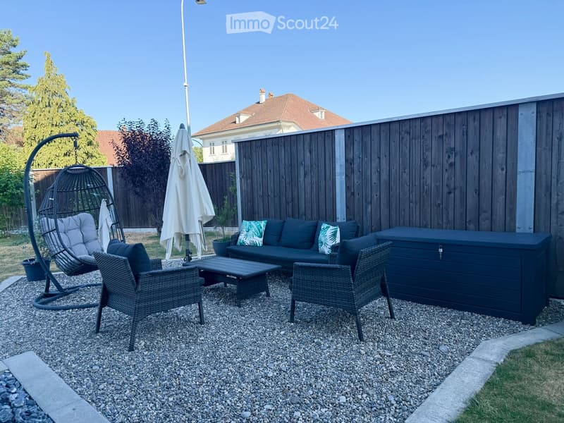 Garden area with a sofa, coffee table, wicker chairs, a hanging egg chair, and an umbrella, surrounded by a wooden fence and greenery.