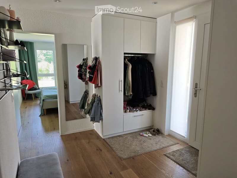 A well-lit hallway with a large white wardrobe, a mirror, a bed in the background, and a shelf with folded clothes. Shoes are placed on the floor near the entrance.