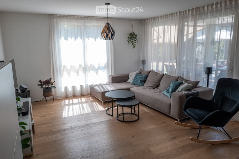 Modern living room with wooden flooring, large windows with sheer curtains, a grey sectional sofa with multiple cushions, a black coffee table, a rocking chair, and a pendant light fixture.