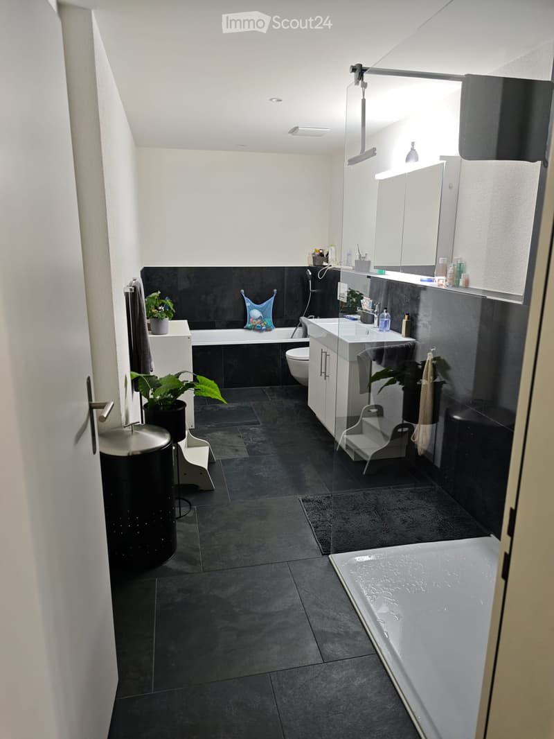 Modern bathroom with dark tiled floor and walls, featuring a bathtub, sink, toilet, mirror, and small stool. Includes plants and towels.