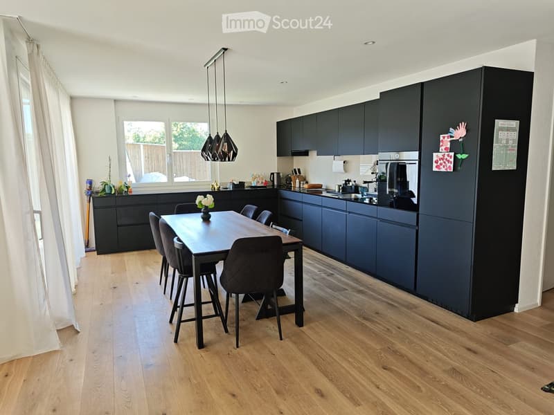 Modern kitchen with dark blue cabinetry, a wooden dining table with six chairs, and various appliances including a coffee maker, refrigerator, and oven. The space features wooden flooring and large windows with white curtains.