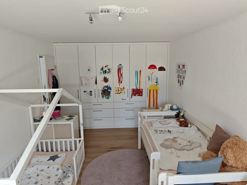 A well-lit children's bedroom with a bunk bed, a small bed, a wardrobe, a desk, and various toys and decorations.