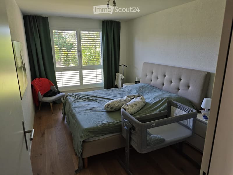 Bedroom with a large bed, nightstand, chair, and a baby cradle. The room features wooden flooring and large windows with blinds.