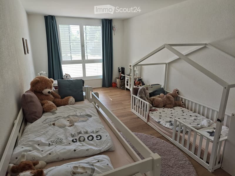 Cozy children's bedroom with a large bed, a small toy house bed, teddy bears, pillows, and a rug on a wooden floor.