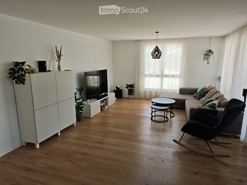 Modern living room with wooden flooring, large windows, a grey sectional sofa, a black rocking chair, a coffee table, a TV, and decorative plants.