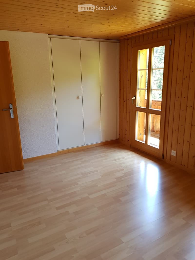 Empty room with wooden flooring, white closet doors, and large windows with wooden frames, allowing natural light to enter.