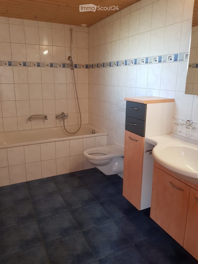 Modern bathroom with a bathtub, toilet, sink, and wooden cabinet. Features tiled walls and dark tiled flooring.