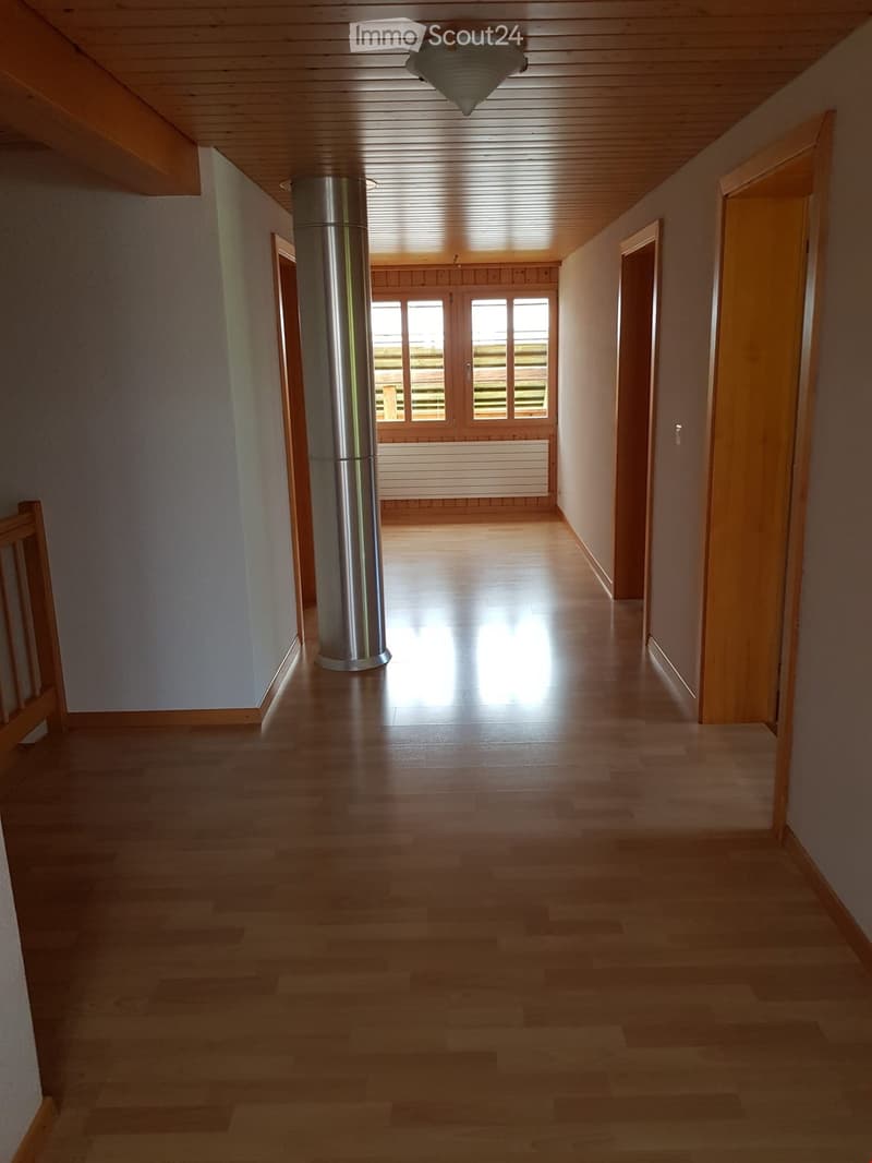 Bright hallway with wooden flooring, light-colored walls, and a central column. Natural light streams in from a large window at the end.
