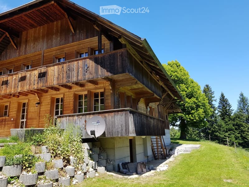 A two-story wooden house with a balcony, satellite dish, and surrounding greenery.