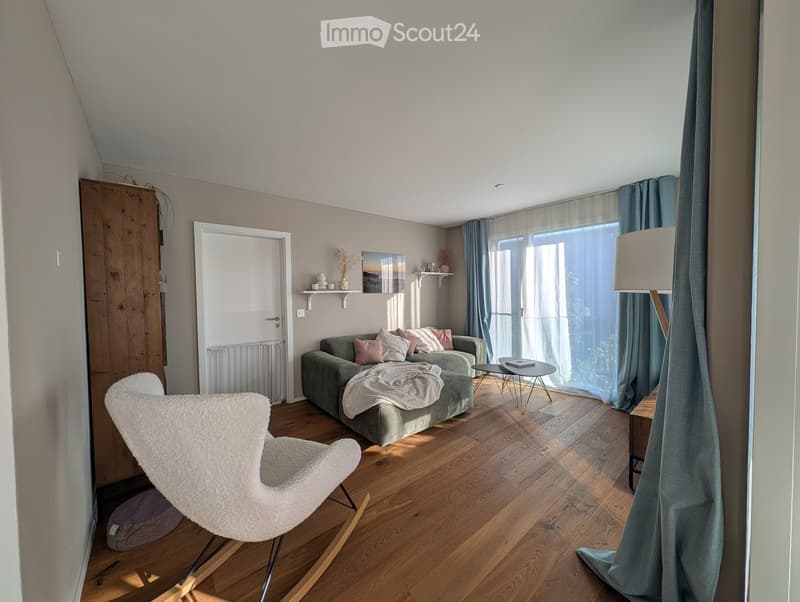Modern living room with wooden flooring, grey sofa, white chair, coffee table, floor lamp, and shelves with decorative items.