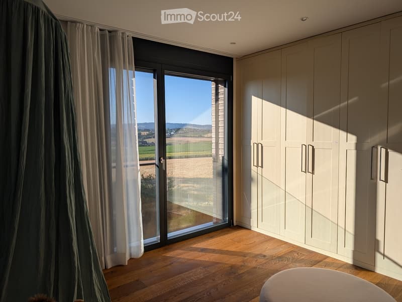 Spacious, well-lit room with wooden flooring, large sliding glass doors leading to a scenic view, built-in wardrobes, and a round stool.