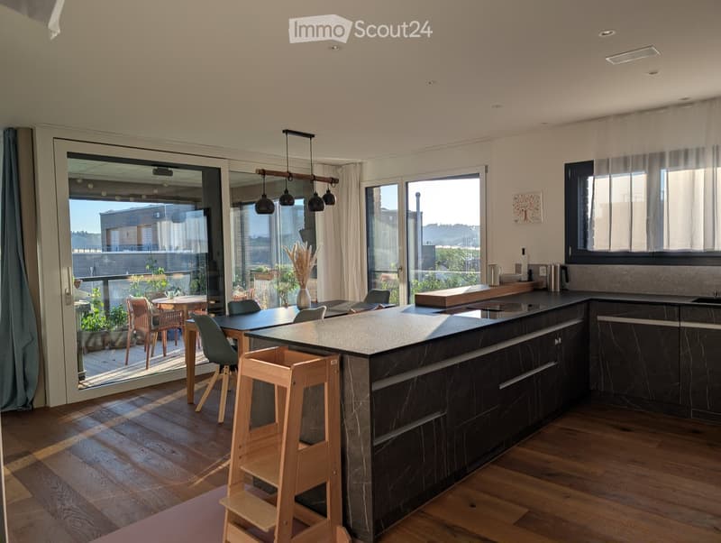 Modern kitchen with dark countertops, wooden flooring, island, bar stools, dining table, and large windows offering a view of the outdoor area.