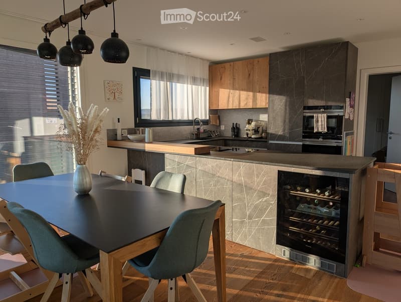 Modern kitchen with a central island, dining table, wine cooler, and various appliances including a coffee machine and oven. The space features wooden flooring, pendant lights, and a large window with blinds.