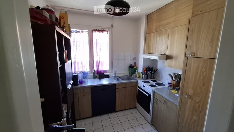 A small kitchen with wooden cabinets, a sink, an oven, and various kitchen utensils on the counter. There is a refrigerator on the left side and a window with curtains above the sink.