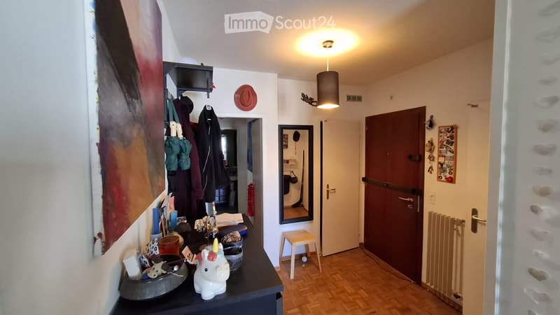 The image shows a well-lit hallway with a wooden floor, a coat rack with jackets and hats, a small table with various items including a unicorn figurine, a mirror, a stool, and several framed pictures and artwork on the walls.