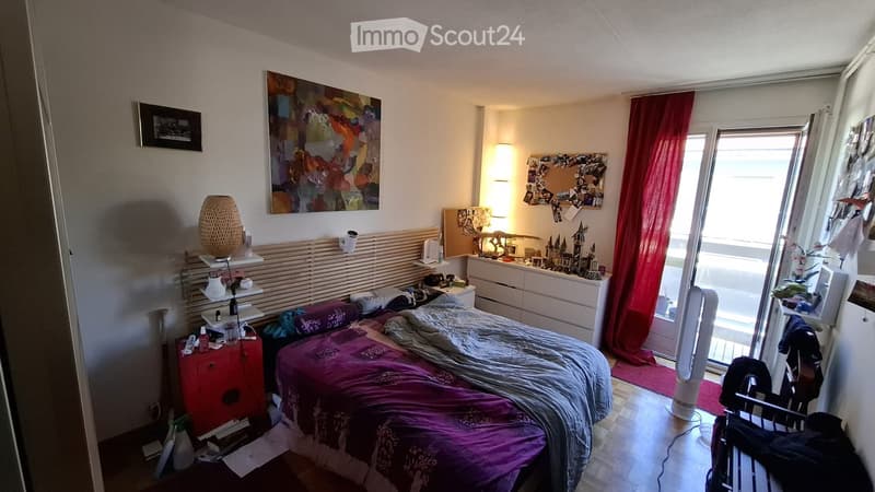 A cozy bedroom with a double bed, nightstand, lamp, and a large window with red curtains. The room features a colorful painting on the wall and a small shelf with decorative items.