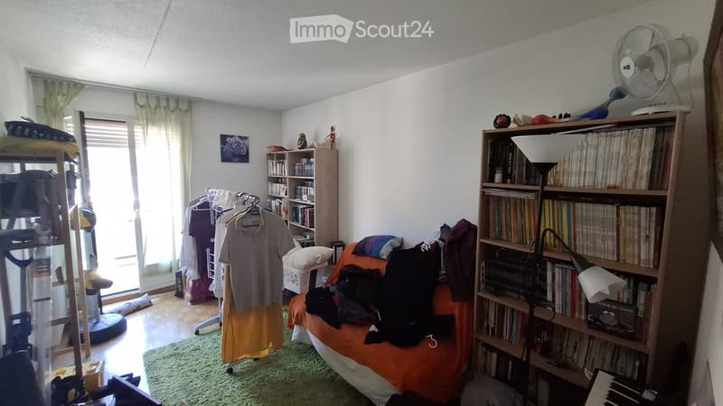 A cozy bedroom with a bed covered in an orange blanket, a bookshelf filled with books, a fan on top, a clothing rack with hanging clothes, and a mirror on the wall.