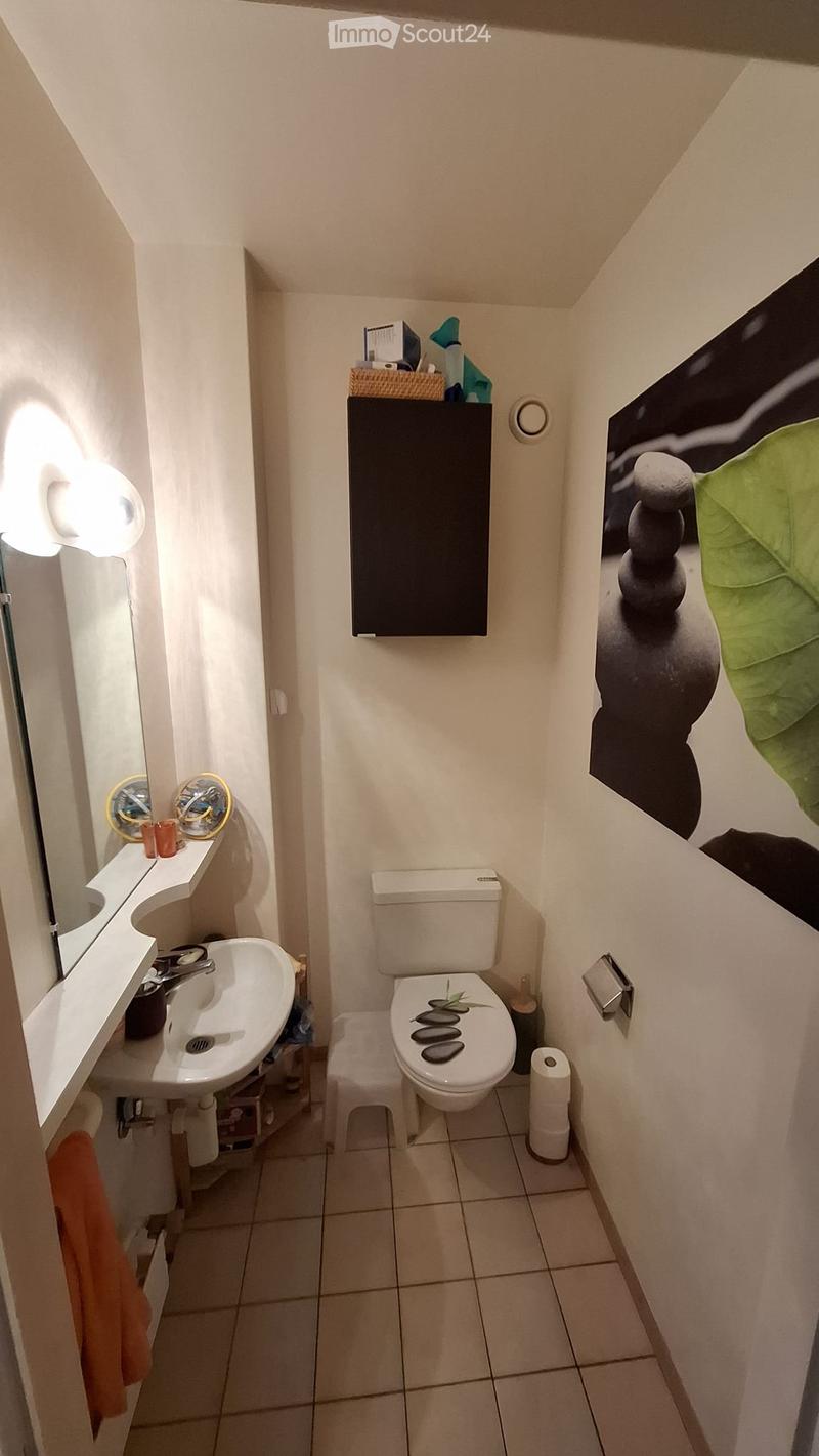 Compact bathroom with white tiled floor, toilet, sink, mirror, and a small stool. Features a decorative picture frame and a cabinet with a basket on top.