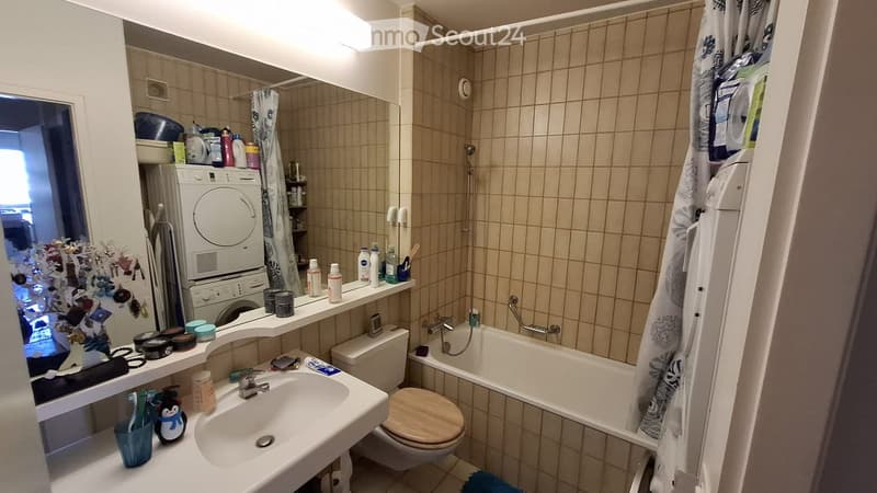 Bathroom with beige tiled walls, a sink, toilet, bathtub, washing machine, and various toiletries on shelves and counters.