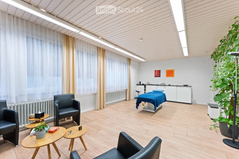 Modern, well-lit room with light wooden flooring, white walls, and large windows covered with sheer curtains. Features include a massage table, two black leather chairs, small wooden tables, a potted plant, a tall floor lamp, and a white cabinet with a co