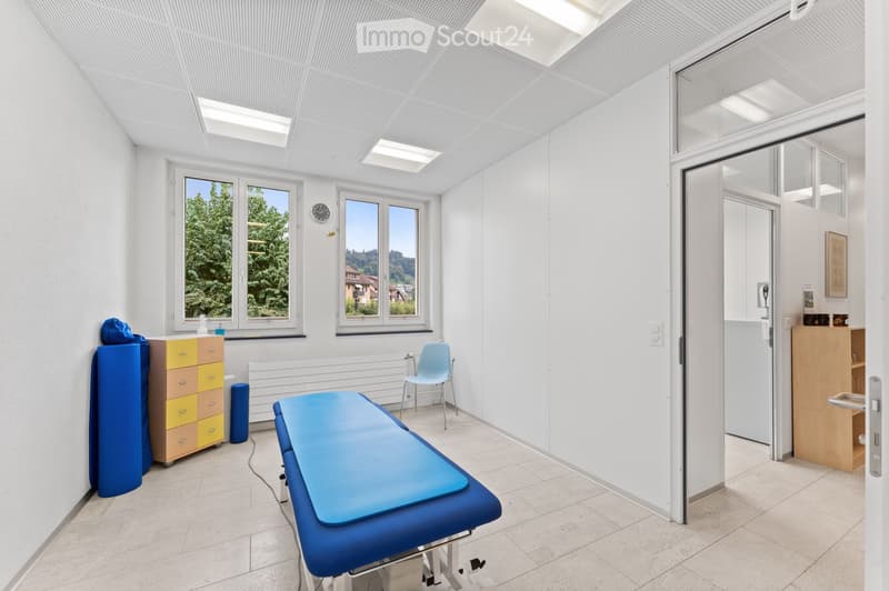 A clean, well-lit examination room with a blue examination table, a light blue chair, a yellow and brown cabinet, and a clock on the wall. Large windows offer a view of greenery outside.