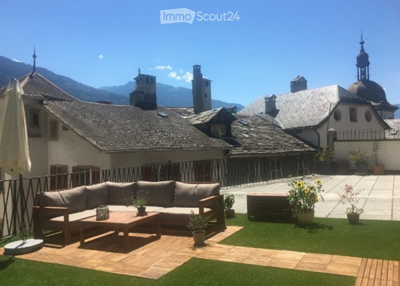 Balcony or terrace area with artificial grass, wooden decking, grey sectional sofa, coffee table, potted plants, and an umbrella.