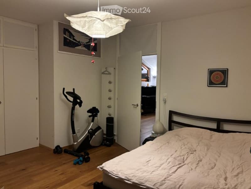 A bedroom with a bed, exercise bike, and a picture frame on the wall. The room has wooden flooring and a hanging lamp.