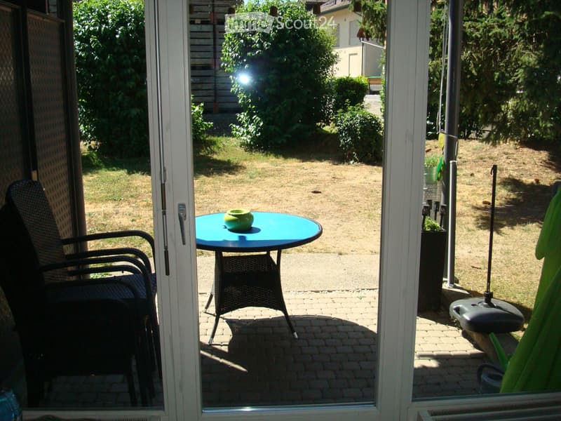 Balcony with a blue round table, black wicker chair, and a green vase on the table. The balcony overlooks a grassy yard with trees and a house in the background.
