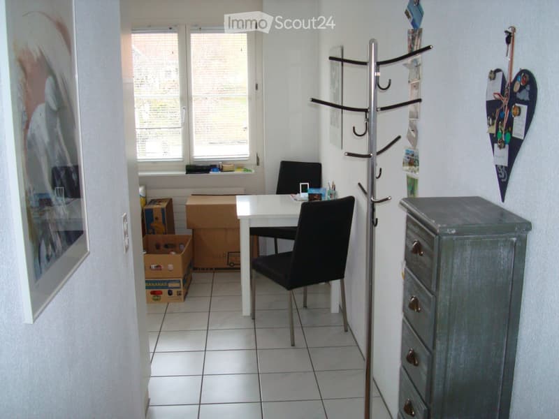 A small, furnished room with a white table, black chair, grey cabinet, coat rack, and several cardboard boxes near the window.