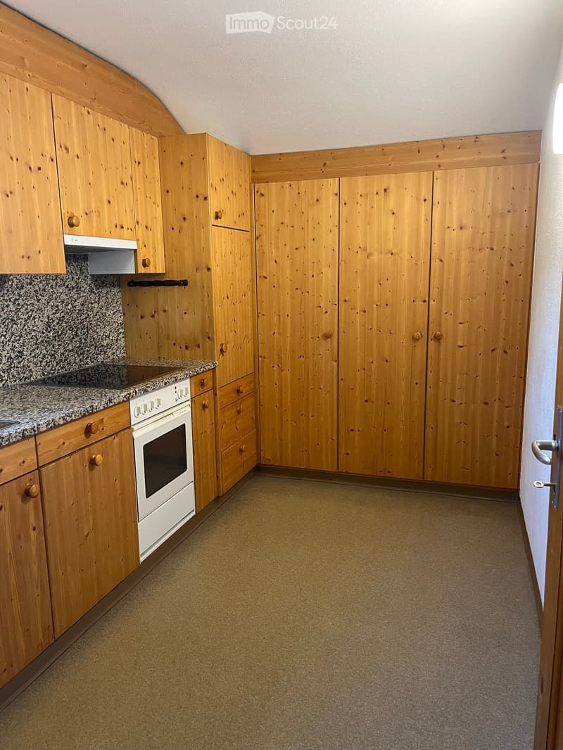 A small kitchen with wooden cabinets, a white oven, and a granite countertop. The floor is carpeted, and the walls are painted white with wooden paneling above the cabinets.