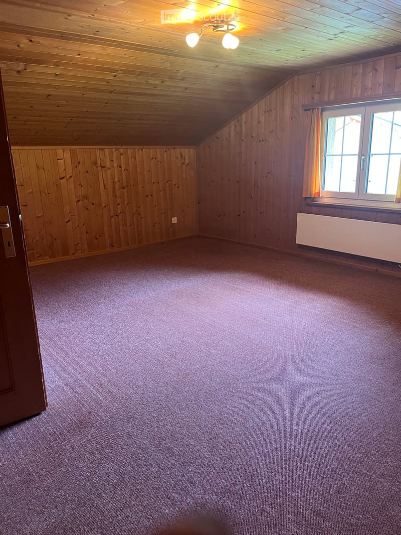 Empty attic room with wooden walls and ceiling, carpeted floor, single window with curtains, and a radiator below it.