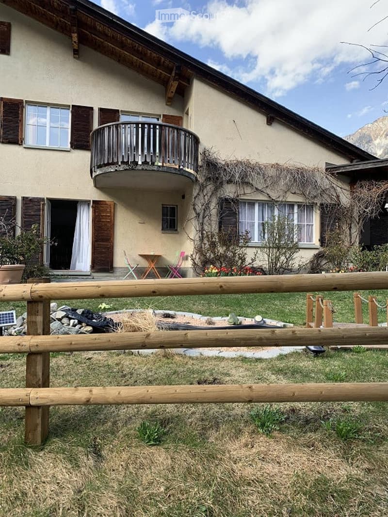 Two-story house with a balcony, wooden shutters, and a small garden area with a table and chairs.