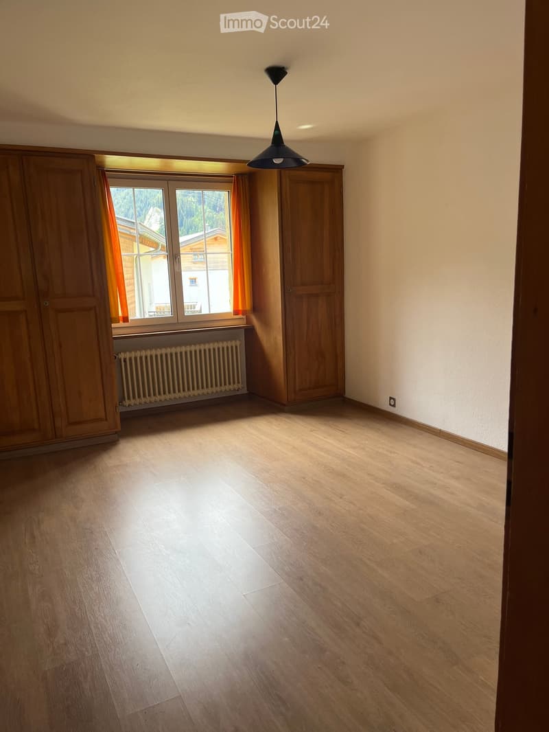 Empty room with wooden flooring, large window with curtains, radiator, and a hanging light fixture.
