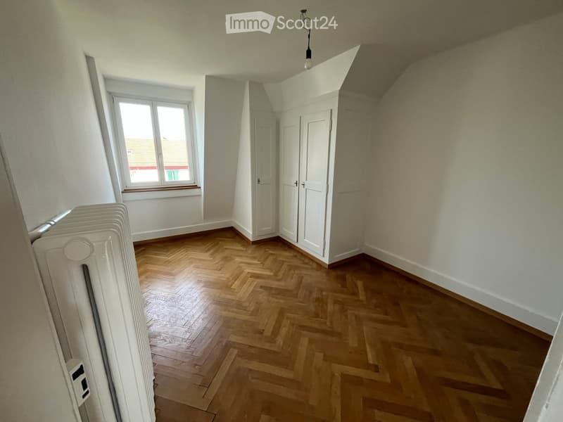 Empty room with wooden herringbone flooring, white walls, and a window with a view of a rooftop area. The room features built-in white closet doors and a radiator on the left side.