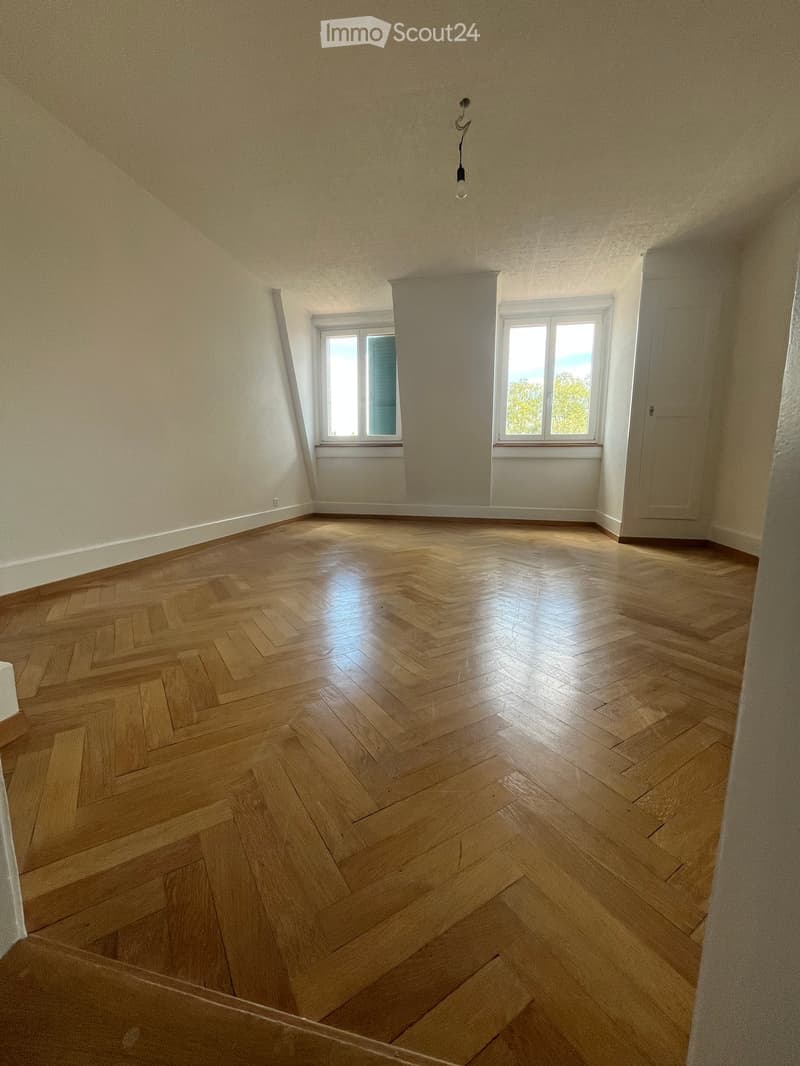 Empty room with wooden herringbone flooring, white walls, and two windows letting in natural light.