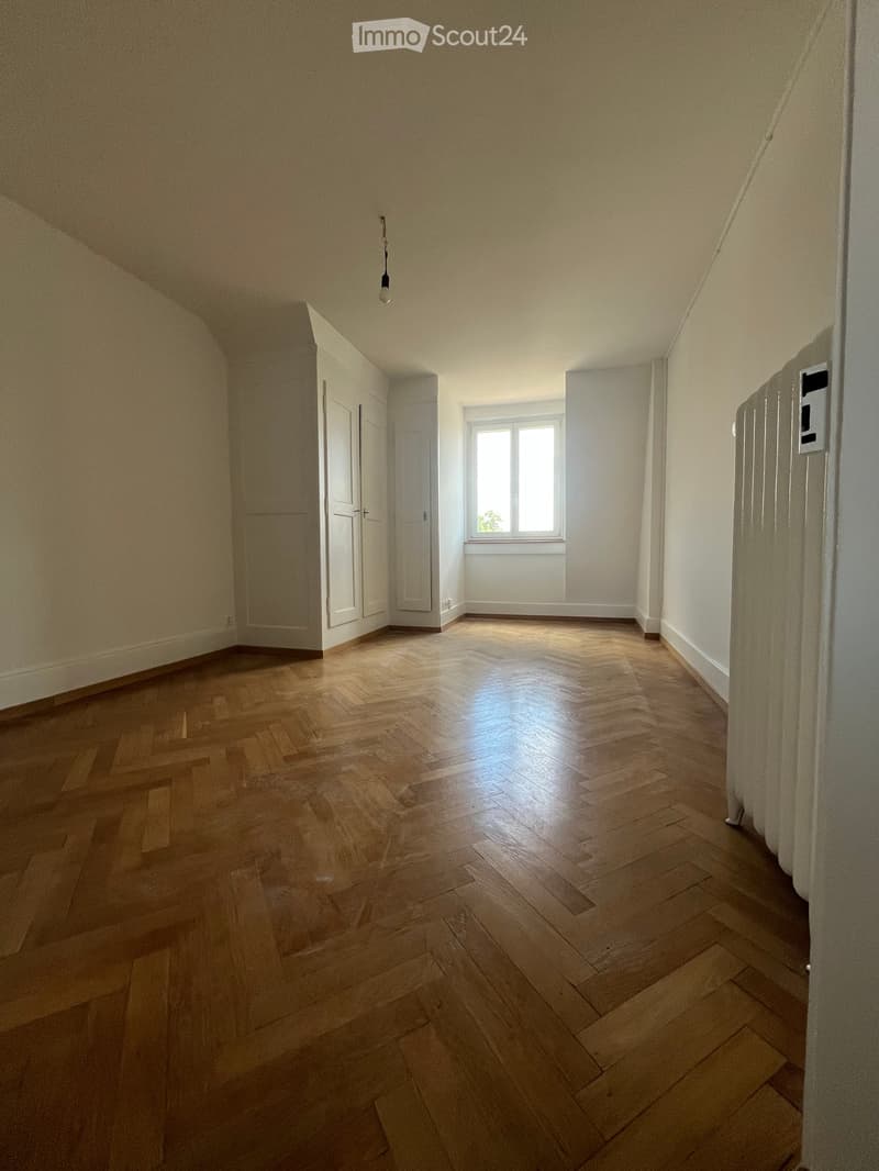 Empty room with wooden herringbone flooring, white walls, and a single window letting in natural light. The room has a door on one side and a radiator on the adjacent wall.