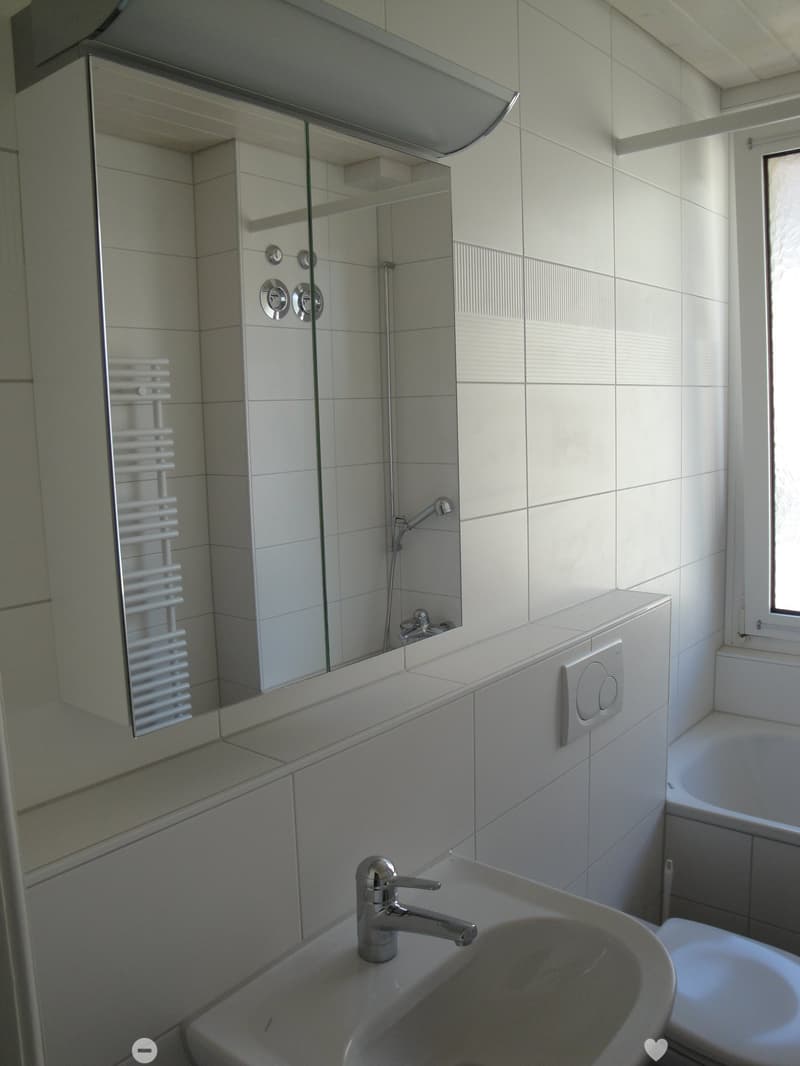 Modern bathroom with white tiles, a sink with a chrome faucet, a large mirror with built-in shelves, a bathtub, and a radiator.