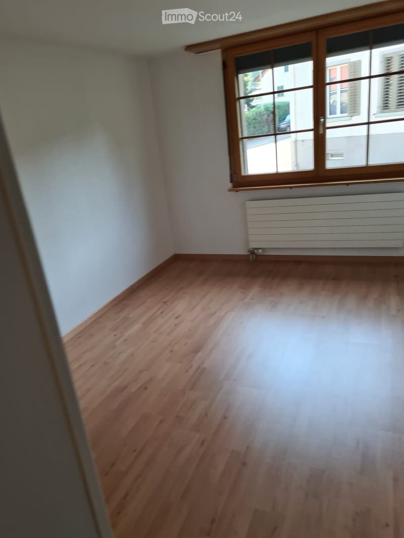 Empty room with light wooden flooring, white walls, and a large window with wooden frame. A white radiator is visible under the window.