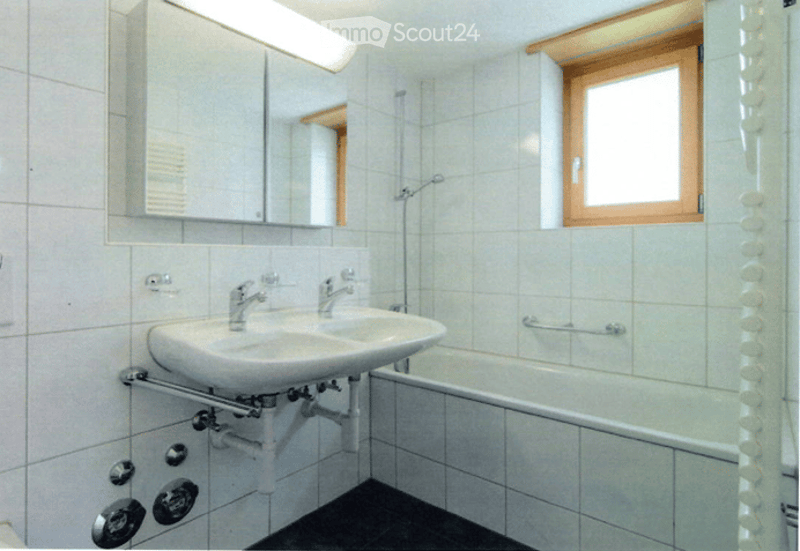 Bathroom with white tiled walls and floor, double sink, mirror, bathtub, and a window with wooden frame.