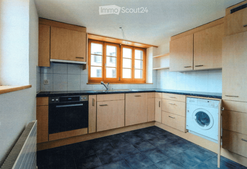 Bright kitchen with wooden cabinets, a window above the sink, and a washing machine. The floor is dark blue tiles, and the walls are white with blue tiles around the sink area.