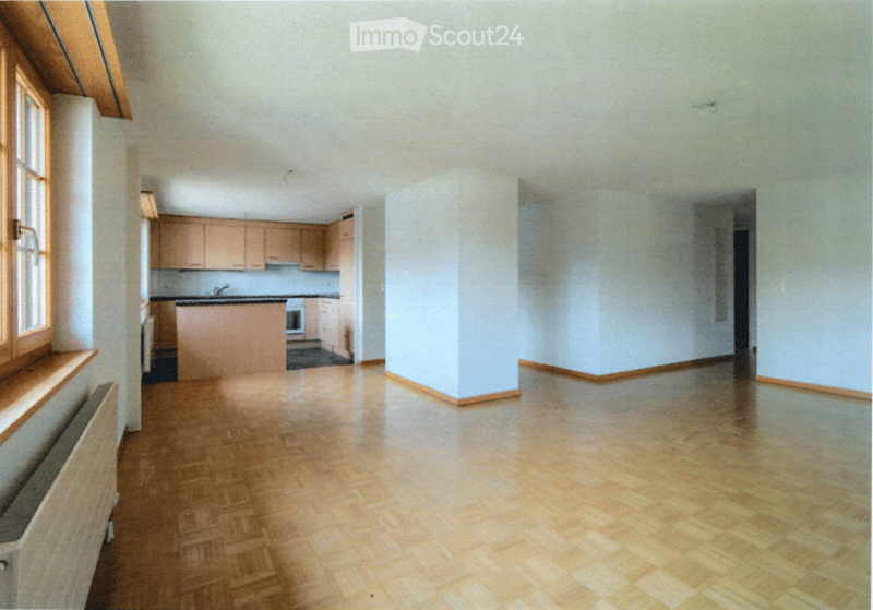 Empty room with wooden flooring, white walls, and a small kitchen area visible in the background. No furniture or equipment present.