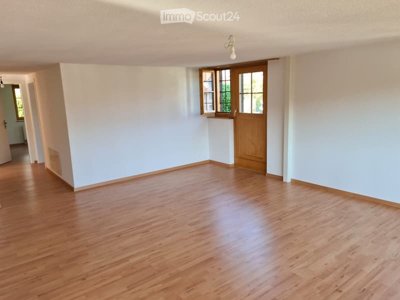 Empty room with light wooden flooring, white walls, and a wooden door leading to another space.