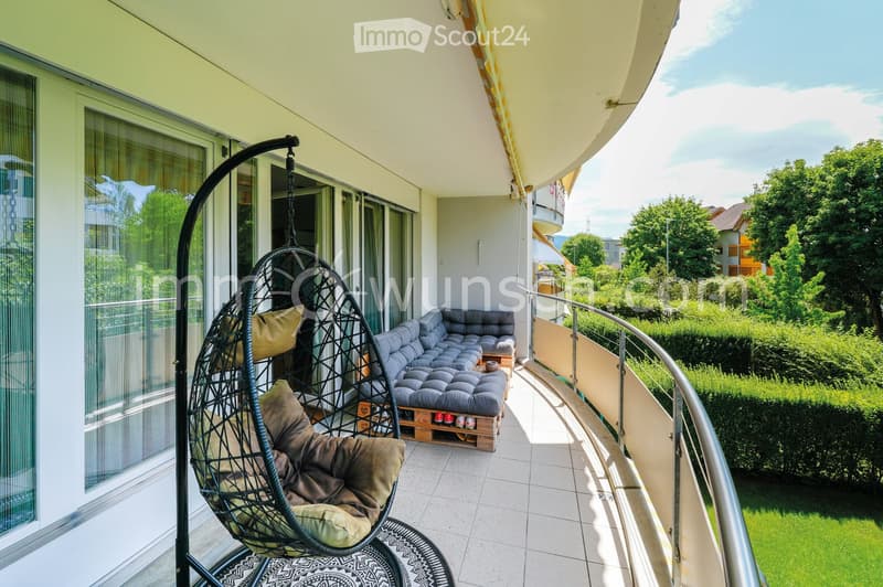 Balcony with a modern design, featuring a grey sectional sofa, a wicker hanging egg chair with cushions, a small wooden table, and a patterned rug.
