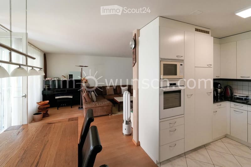 A modern, open-plan living and dining area with a kitchen. The living area features a brown sofa, a coffee table, a piano, and a floor lamp. The dining area has a wooden table with black chairs. The kitchen includes white cabinets, a microwave, an oven, a