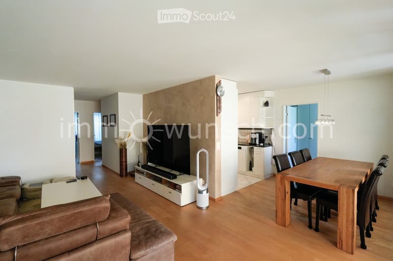 Open-plan living and dining area with a brown leather sofa, coffee table, flat-screen TV, wooden dining table with chairs, and a small kitchen area with cabinets and a coffee machine.