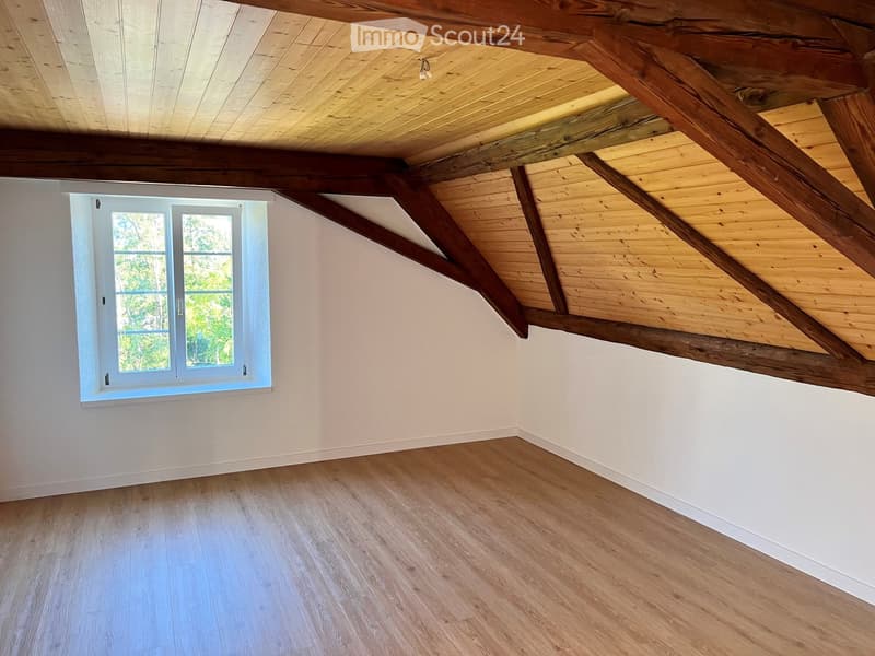 Empty room with wooden floor, white walls, and exposed wooden beams on the ceiling. A single window with white frame allows natural light to enter.