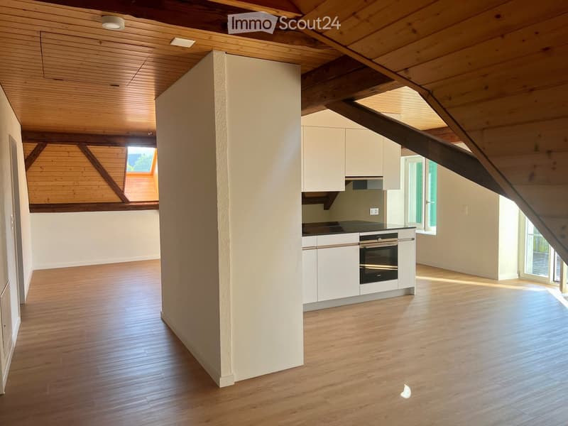 Open-plan attic space with wooden beams, light wood flooring, white cabinetry, and an integrated oven. Skylight and large windows provide natural light.